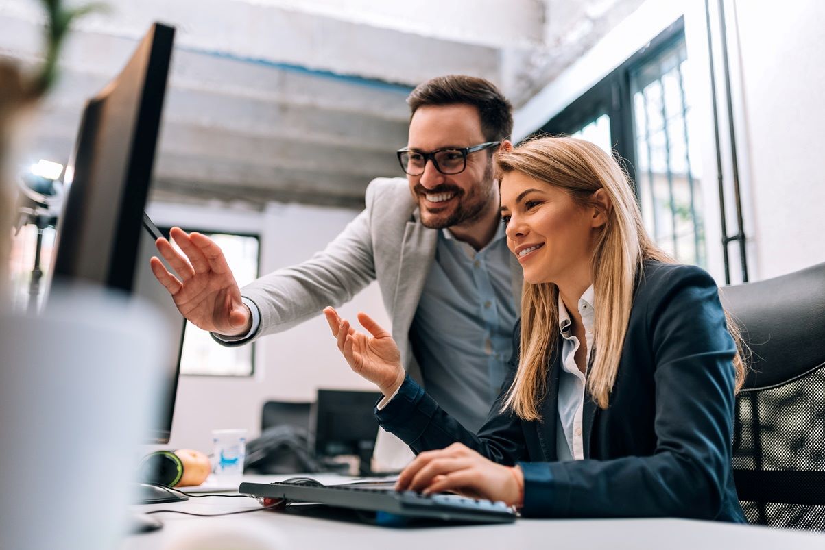 A man and woman in business attire smile as they collaborate at a computer in a bright office. The man gestures at the screen, conveying teamwork.